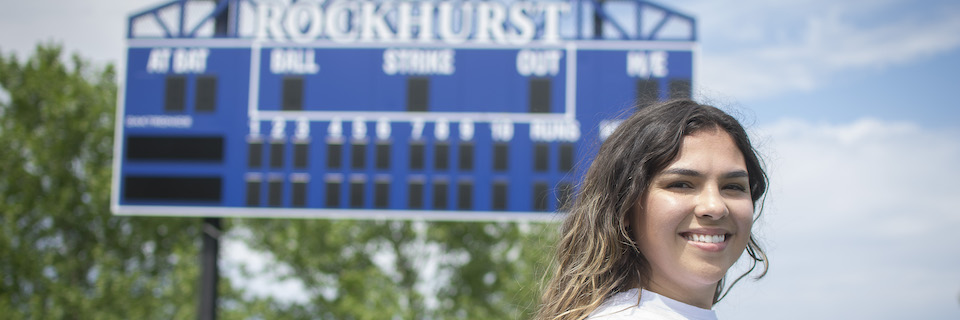 BreAnna Droge standing in front of the softball scoreboard on campus