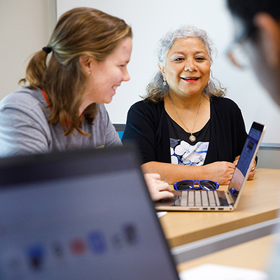 A professor talks with a student working on a laptop