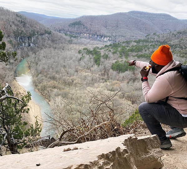 Student Fazion Campbell points across a picturesque view of a river from a cliff while hiking in Arkansas
