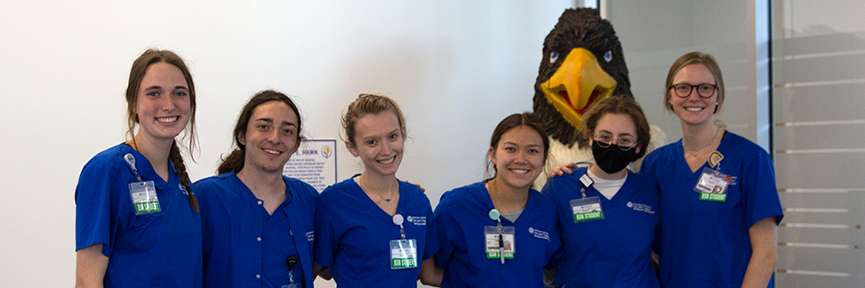 Six nursing students in blue Rockhurst scrubs in front of wood carving of Rock E. Hawk mascot