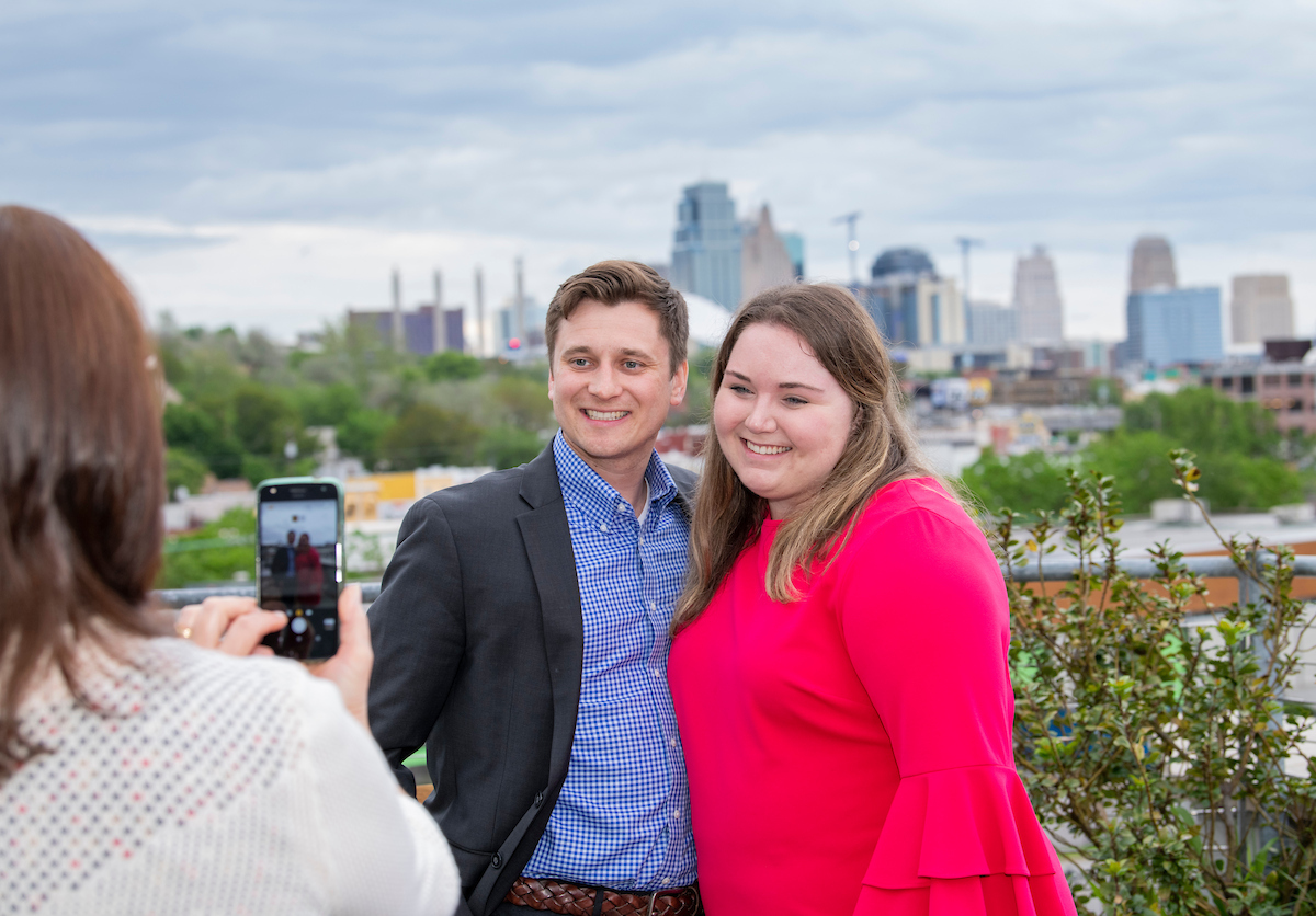 A woman takes a photo of a couple in front of the Kansas City skyline