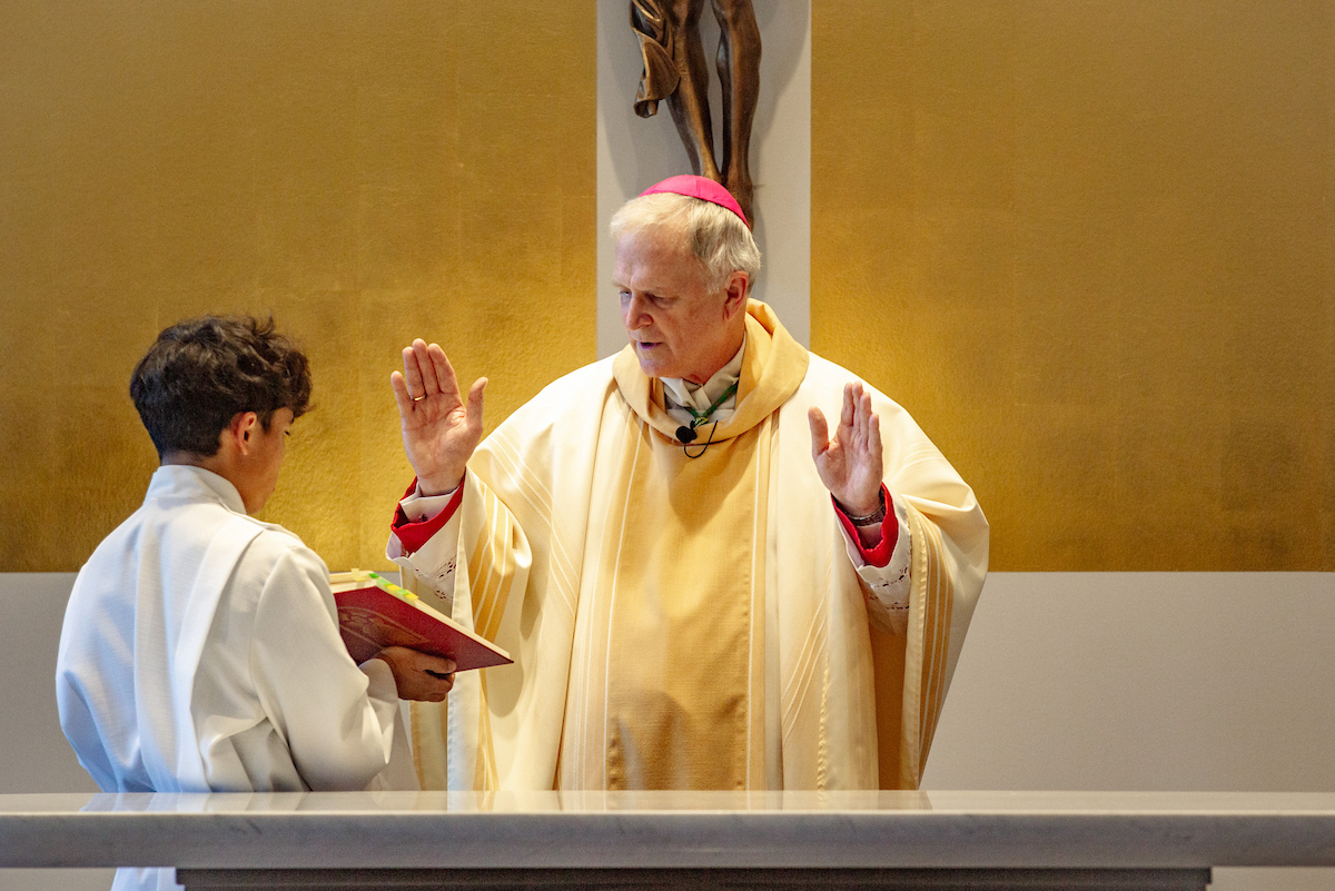 A priest stands with hands raised as he reads from a book held by an altar server
