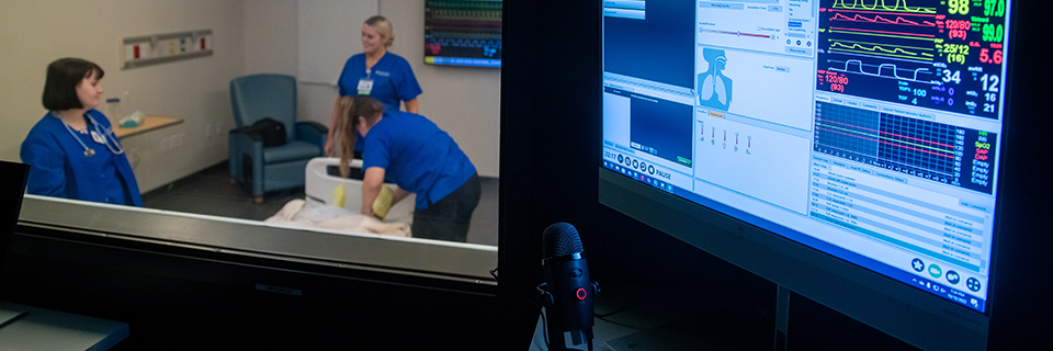 Three nurses in a practicum with vitals monitor in foreground Computer screen in foreground from the observation room overlooking three nurses in the classroom
