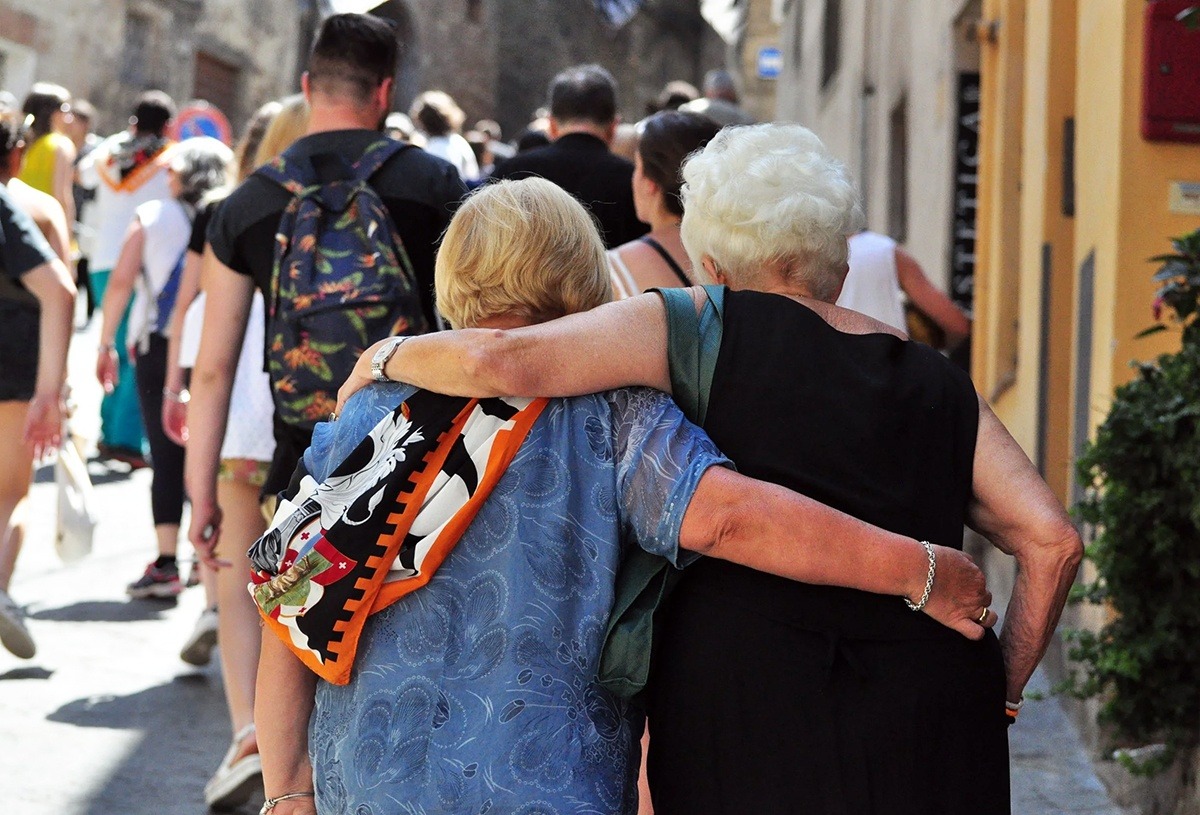 Two women walk on a sidewalk with their arms around each other. Photographed from behind them.