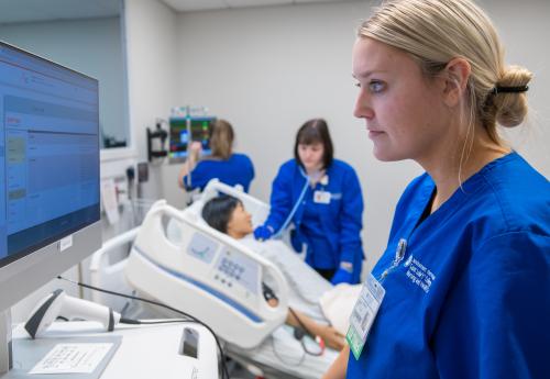 Nursing student at a computer while another checks on a patient and another reads a monitor